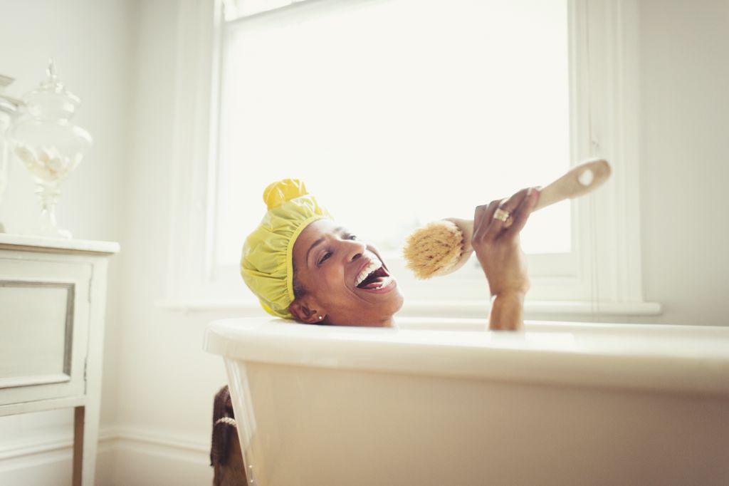 Playful mature woman singing into loofah brush in bathtub