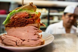 Large meat sandwich with chef in the background; New York City, New York, United States of America