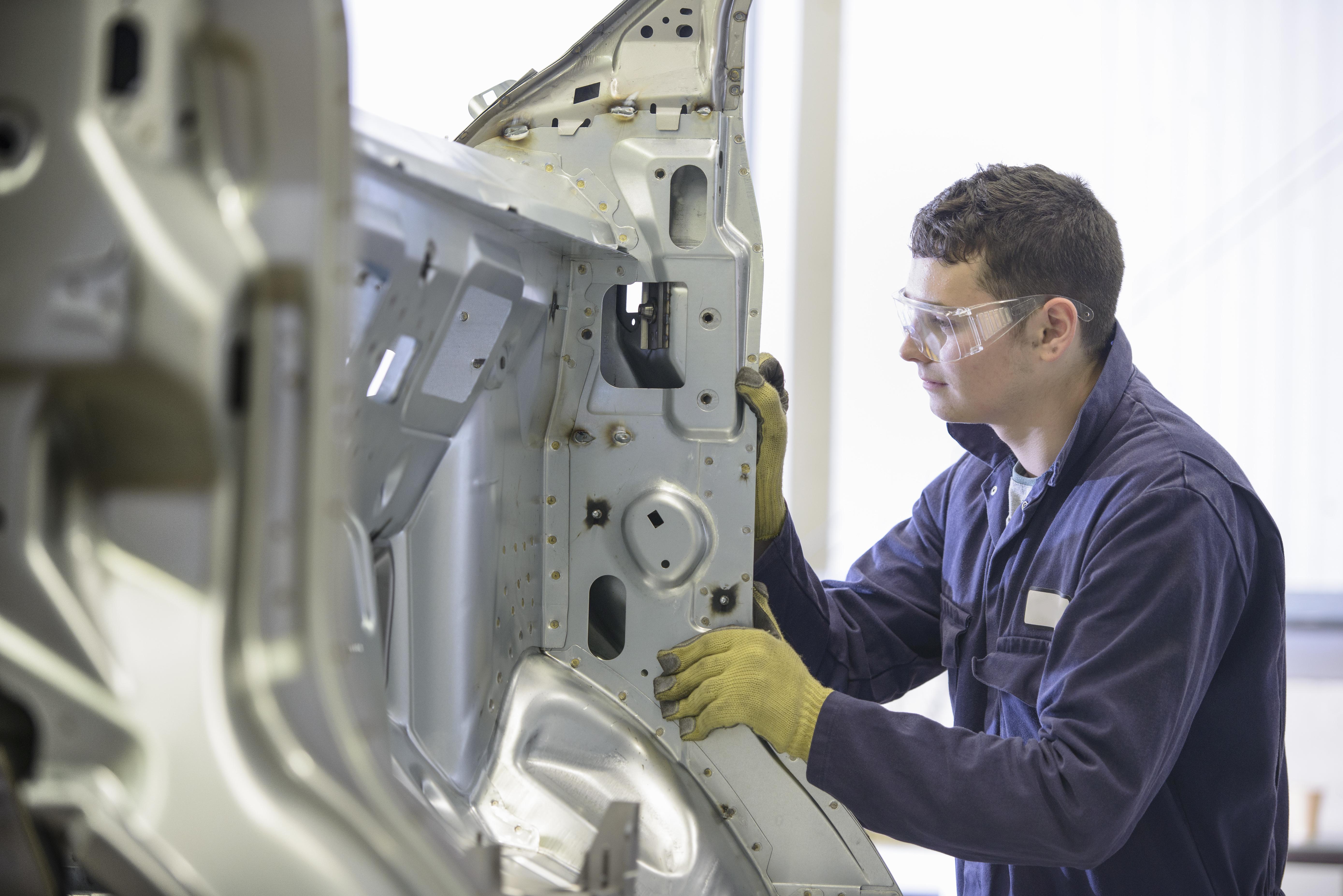 Apprentice wearing boiler suit inspecting car body parts in car plant