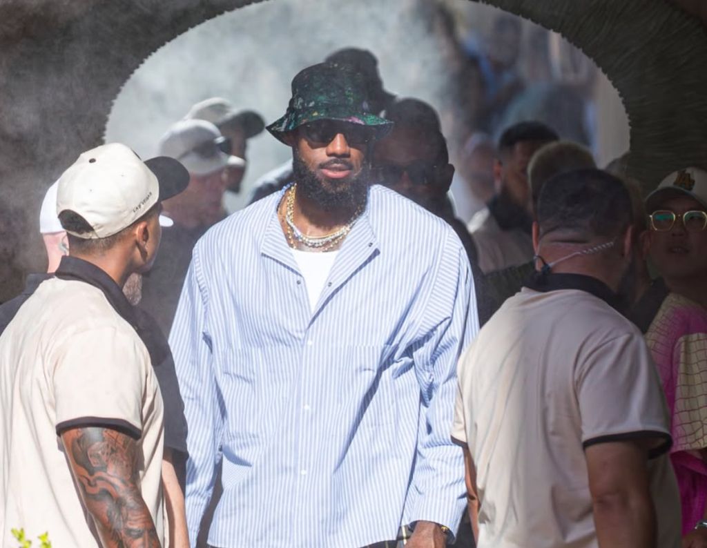 A man wearing a bucket hat and jewelry stands among a group of people in an urban setting.
