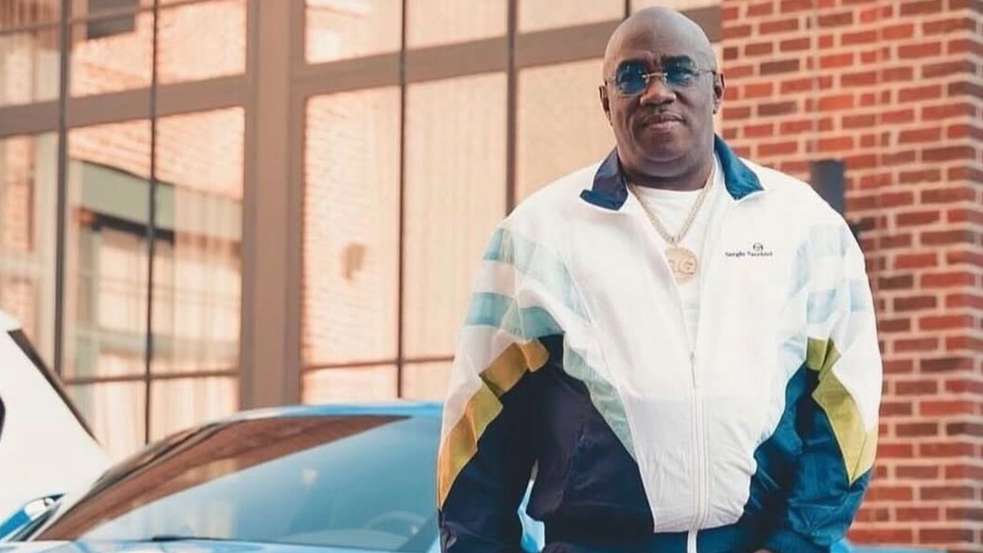 A middle-aged Black man wearing a white jacket with blue and yellow stripes, standing in front of a brick building.