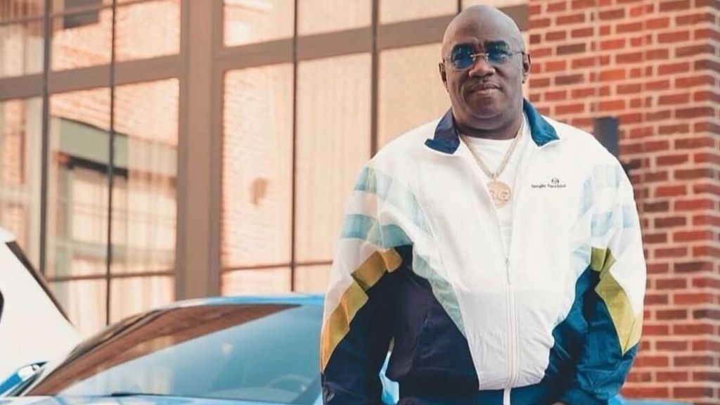 A middle-aged Black man wearing a white jacket with blue and yellow stripes, standing in front of a brick building.