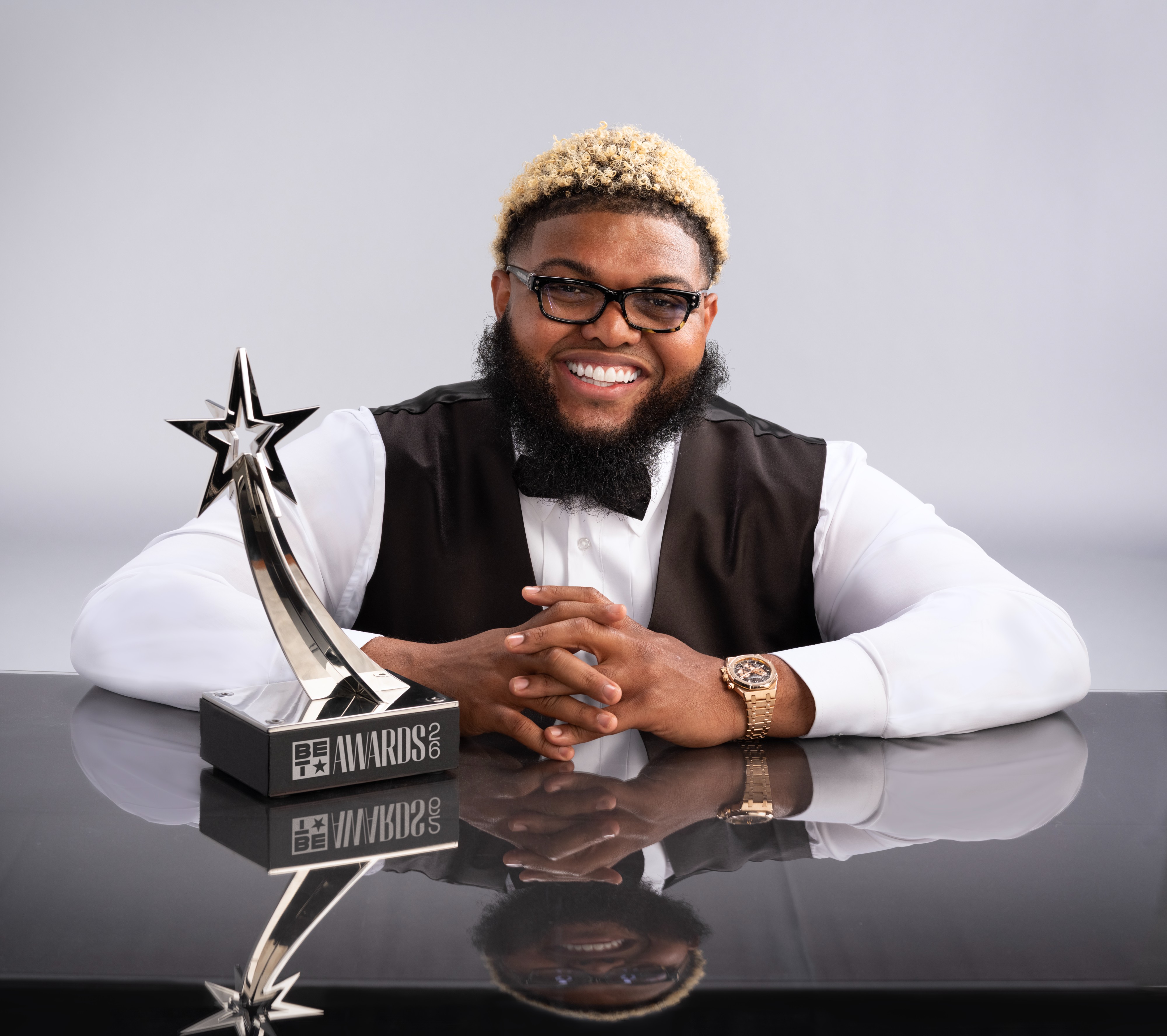 A smiling man wearing glasses and a beard holds a trophy with a star design, seated at a table with a "BET Awards 2022" logo.