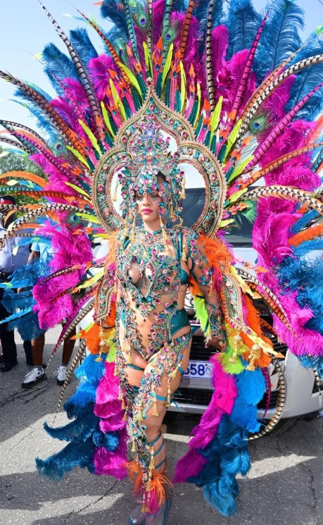 A person wearing an elaborate, colorful feathered costume with a large headdress and holding a mirror, standing in front of a crowd at what appears to be a carnival or festival.