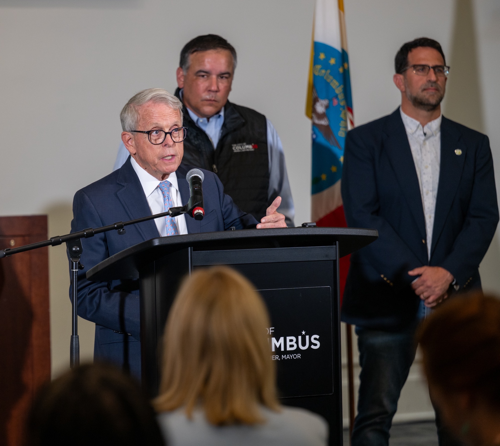 Three men, including a man in a suit speaking at a podium with "MAYOR" text, standing in front of an American flag.
