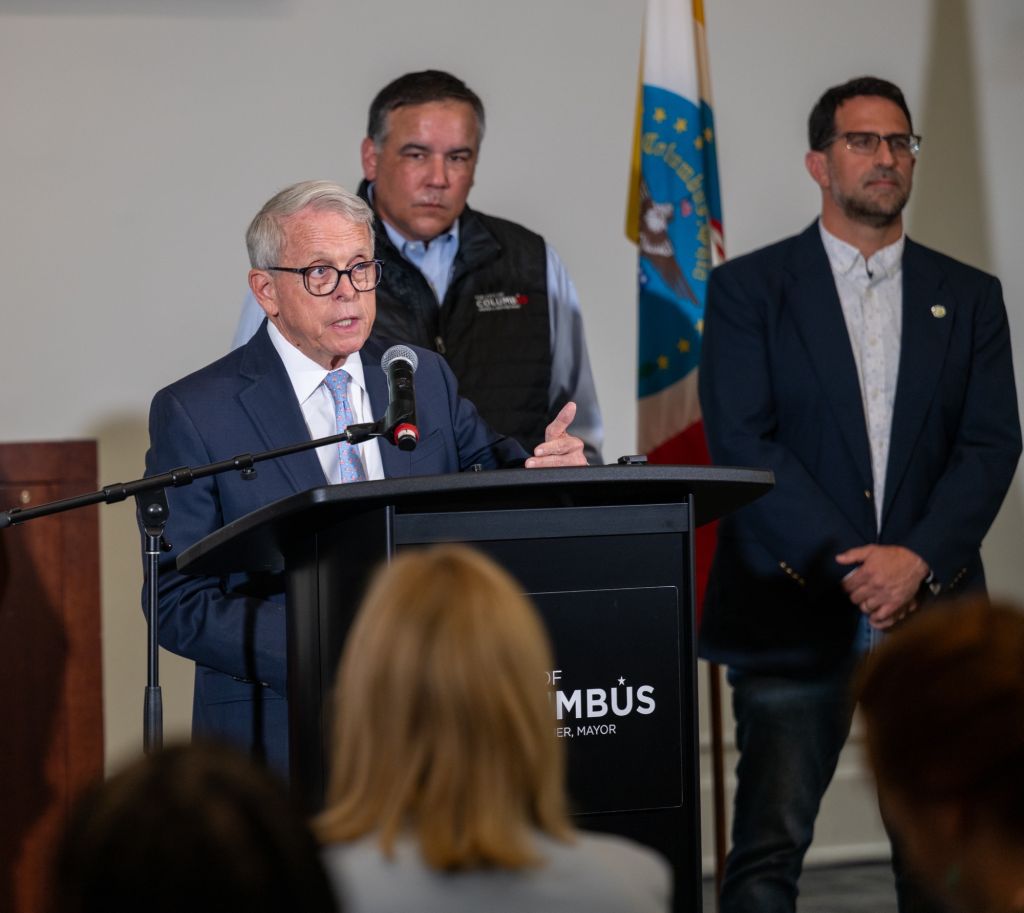 Three men, including a man in a suit speaking at a podium with "MAYOR" text, standing in front of an American flag.